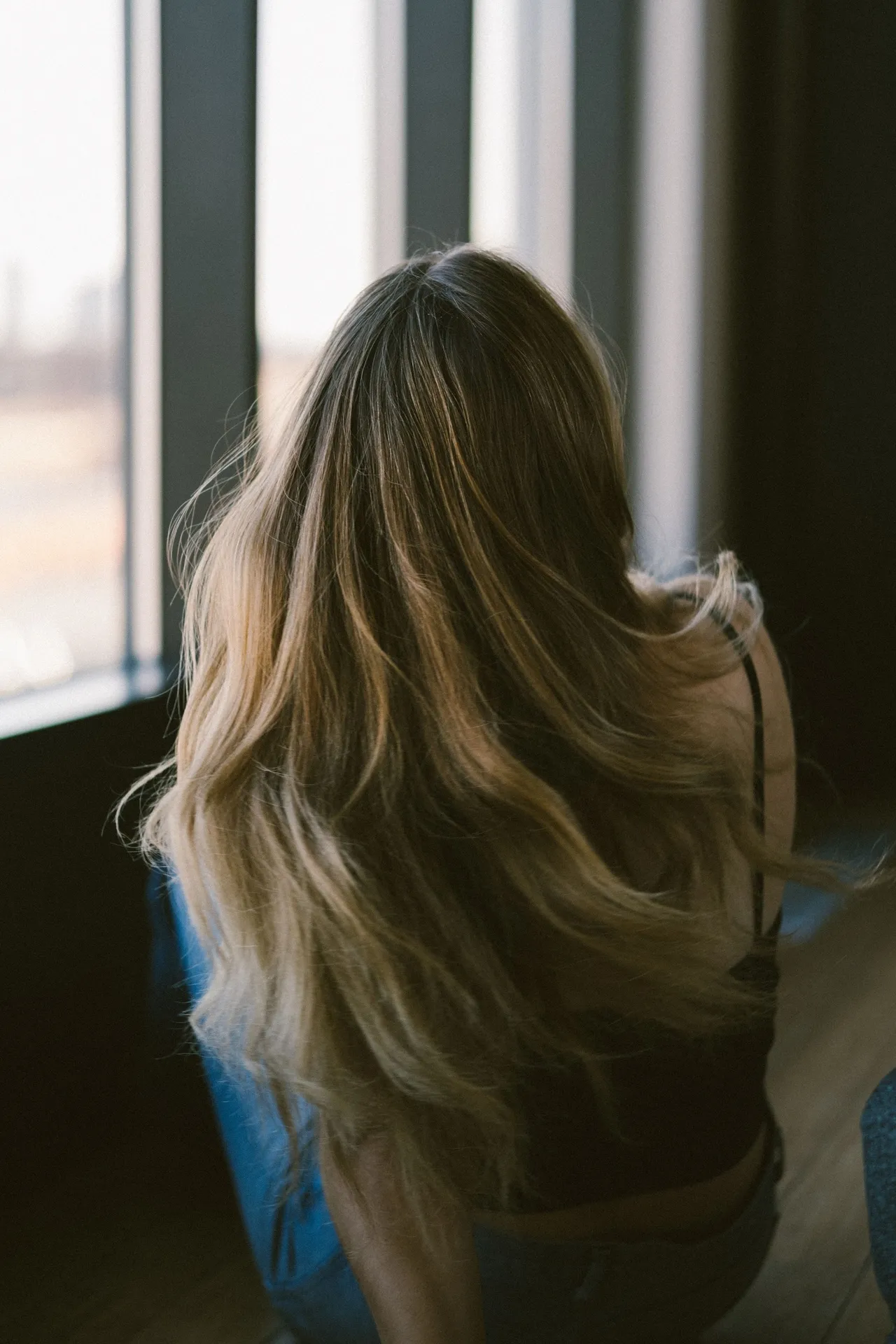 Mulher de costas junto a uma janela, com cabelo comprido iluminado pela luz natural